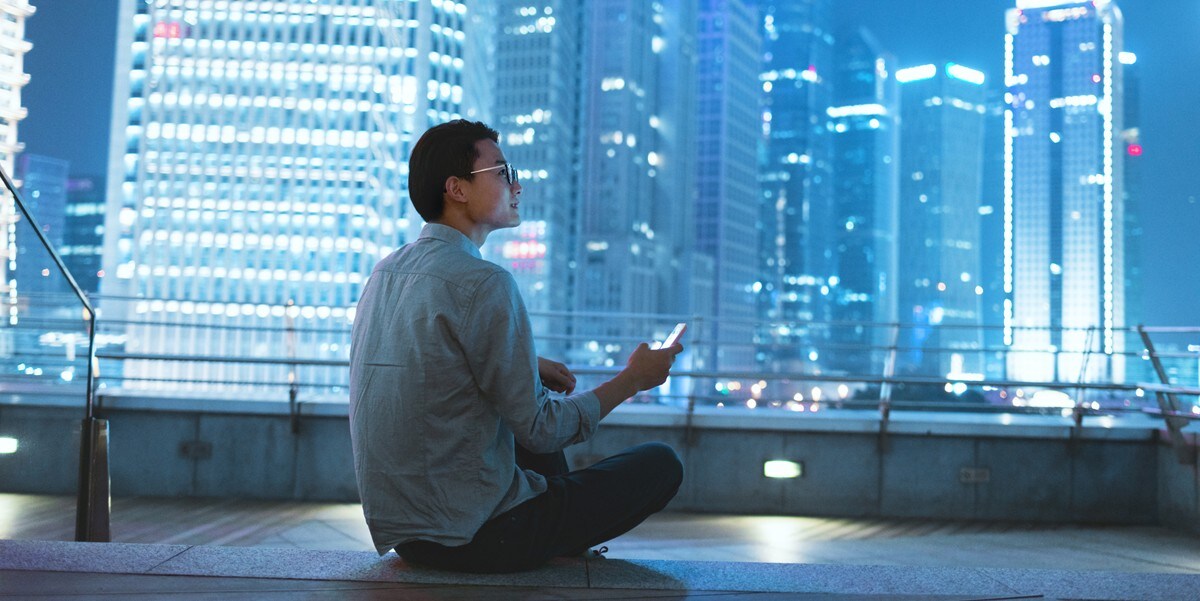 man sitting on rooftop, holding cell phone, lit up skyscrapers in front of him