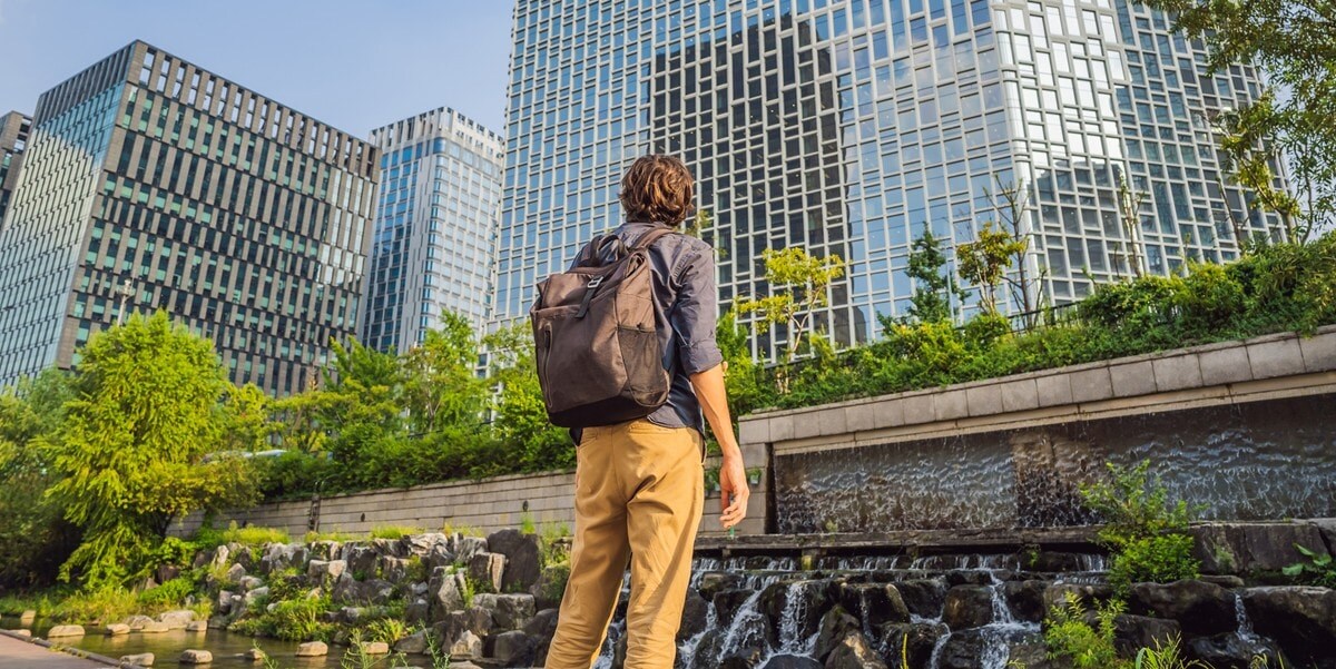 man with backpack standing in park, facing skyscrapers