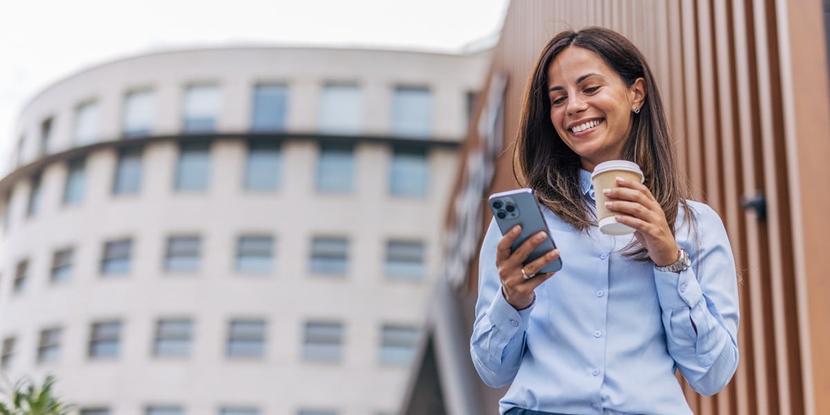 woman standing outside of building with coffee cup in hand, looking at cellphone